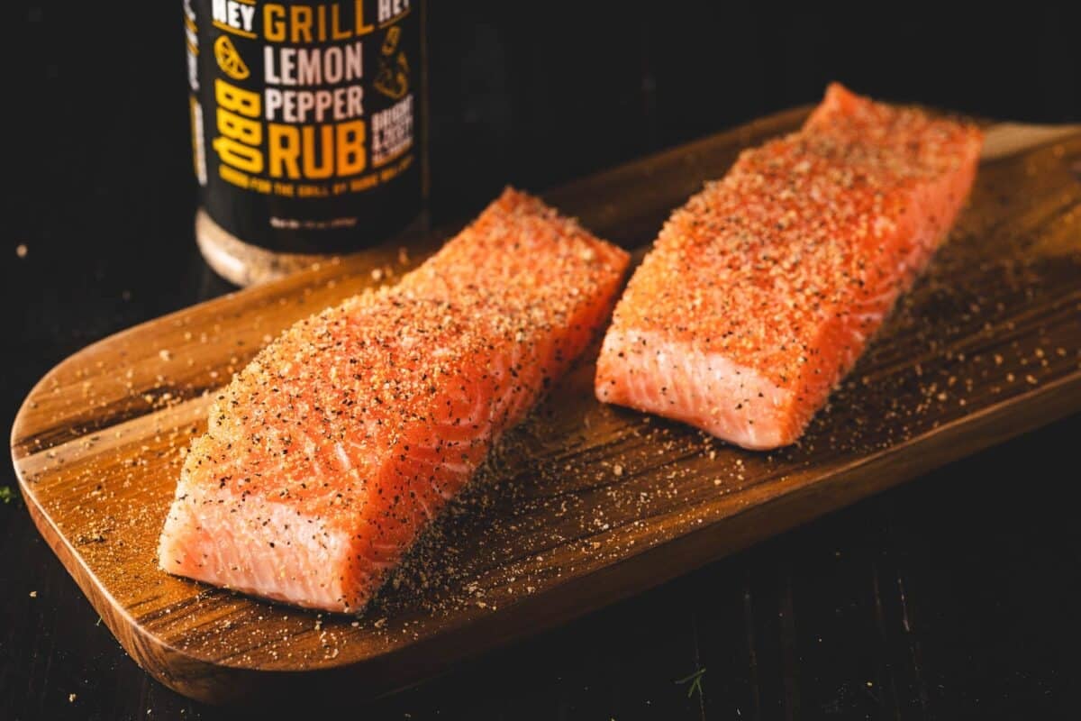 Fish fillets on a wooden cutting board next to a bottle of lemon pepper seasoning.