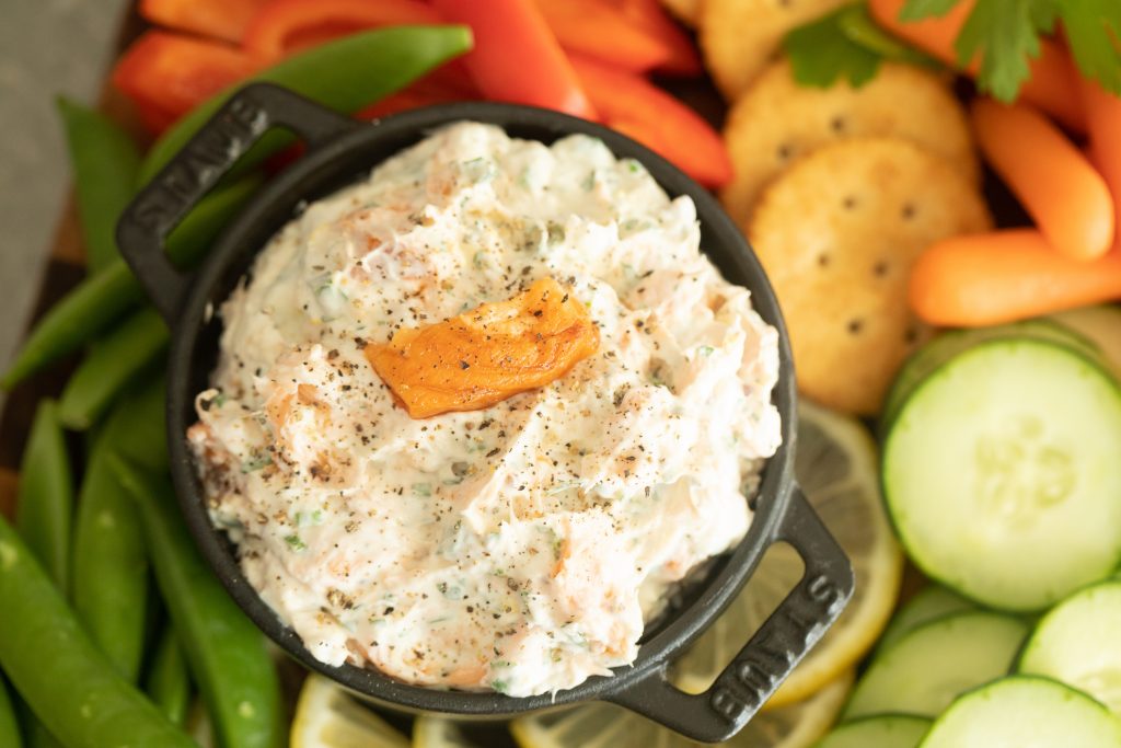 bowl of smoked trout dip in the middle of baby carrots, sliced cucumbers, crackers, snap peas, and sliced red bell peppers.