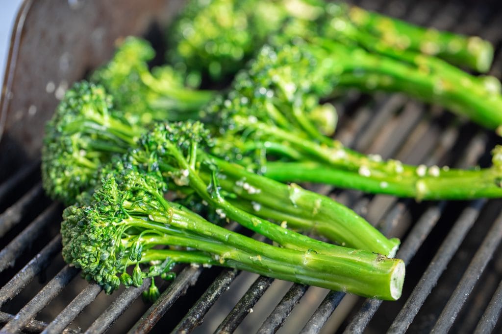 Close up of seasoned broccolini on the grates of a gas grill.