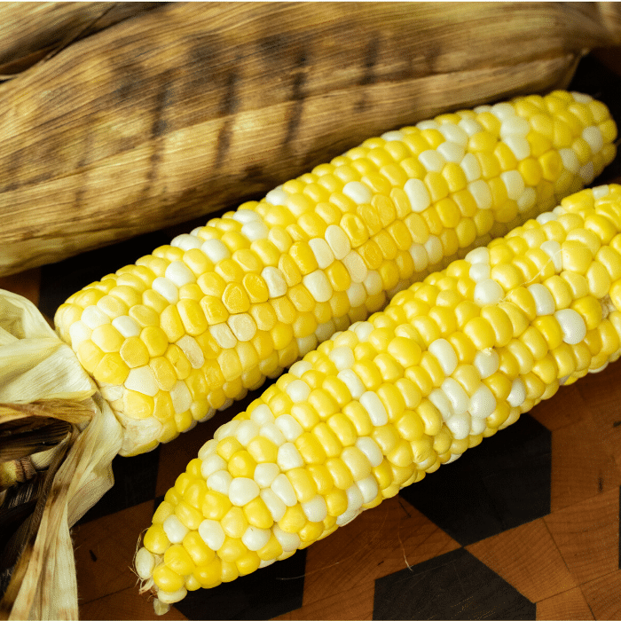 Two grilled and husked ears of corn on a wooden cutting board.