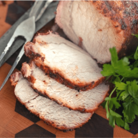 Grilled pork loin sliced into three slices next to a metal fork and knife and fresh herbs on a wooden cutting board.