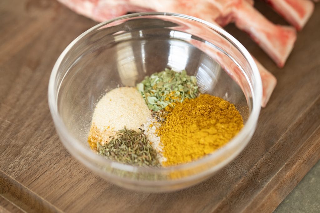 Each of the spices in the recipe unmixed in small glass bowl next to a partially showing raw rack of lamb. All on a wood cutting board.