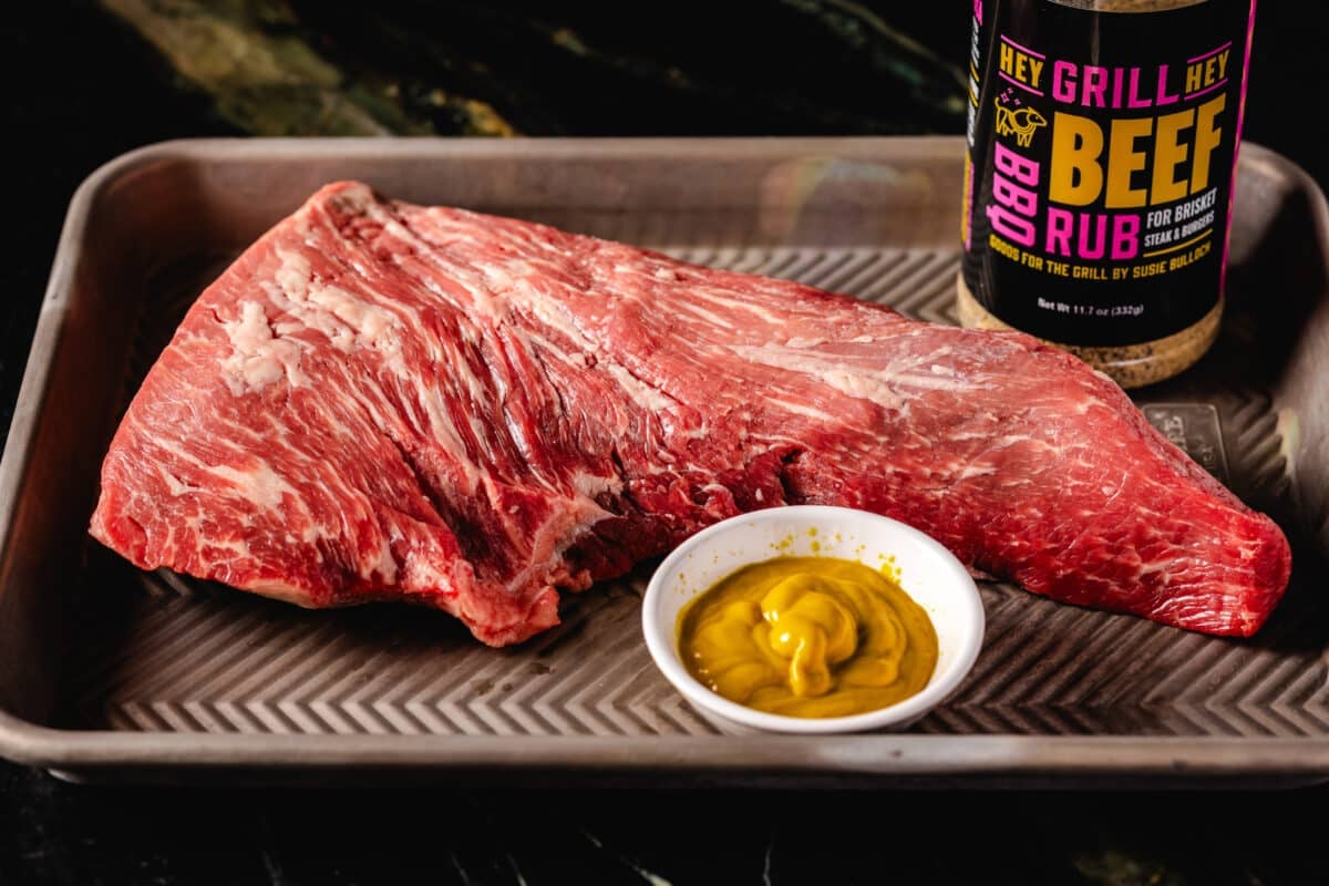 A raw beef roast on a baking sheet with a bowl of sauce and a bottle of Hey Grill Hey Beef Rub.