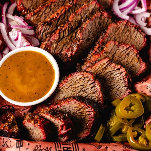 tray of sliced tri tip with a bowl of sauce, sliced red onions, and jalapeño slices.