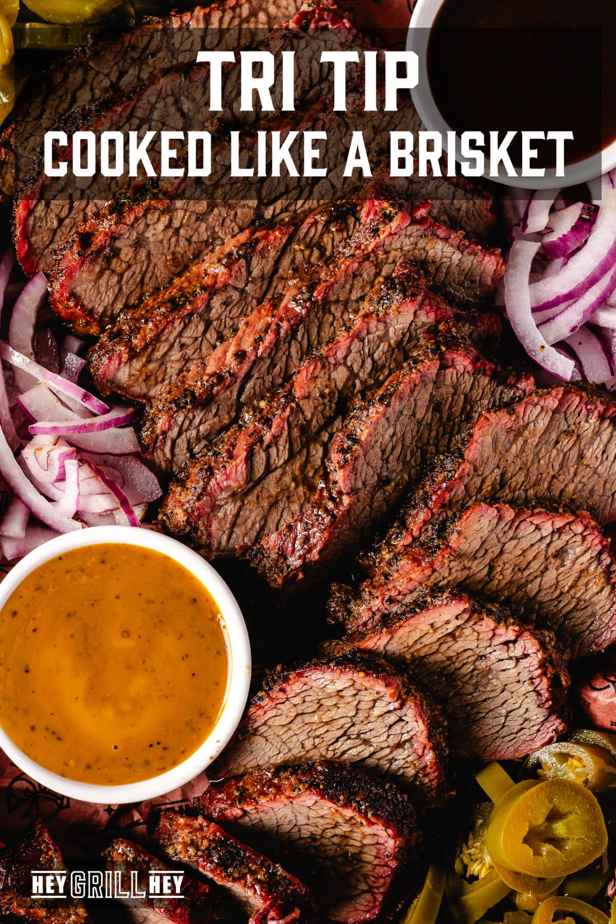 An overhead shot of a tray of sliced tri tip with sauce and condiments. The text overlay reads "Tri Tip Cooked Like a Brisket" at the top, and "Hey Grill Hey" at the bottom.