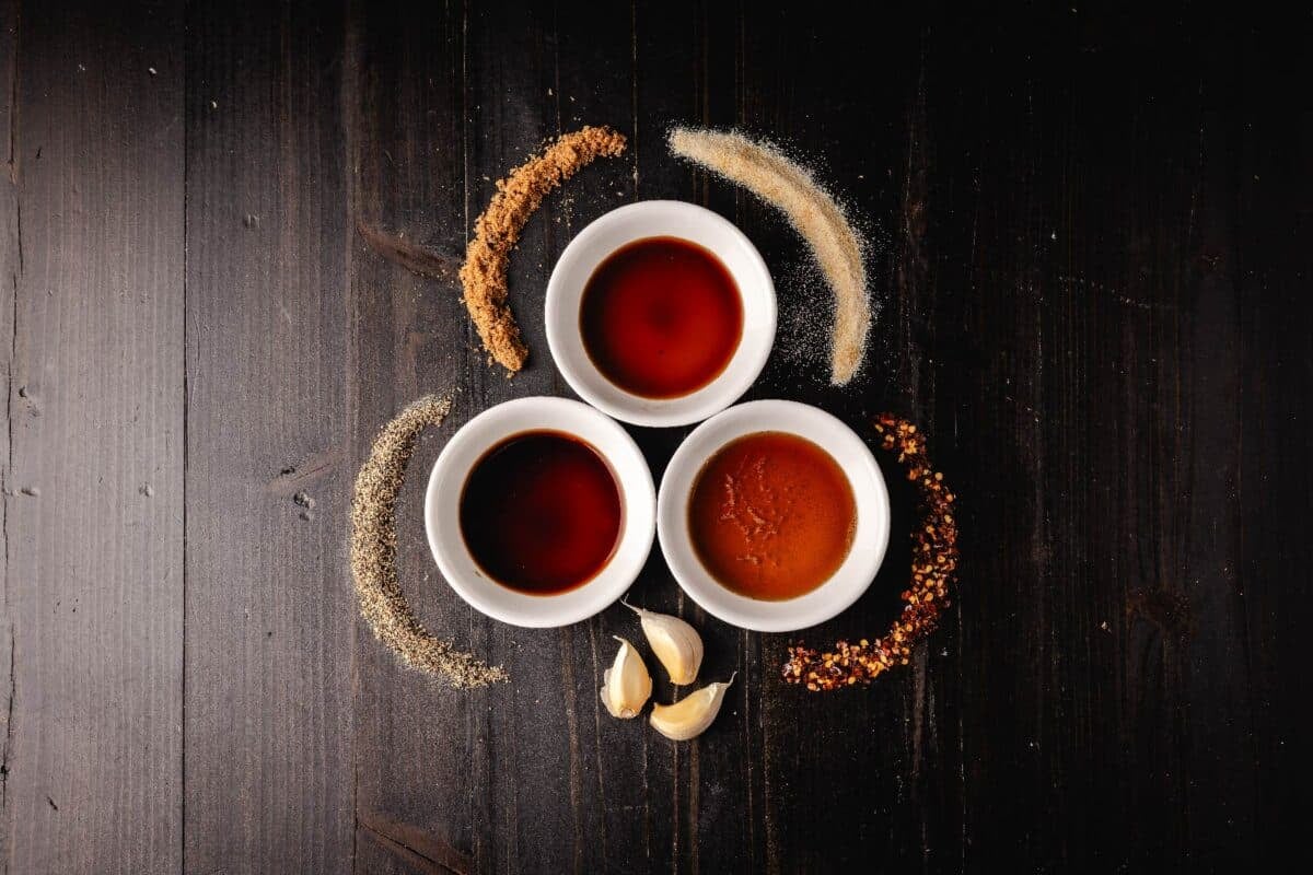 Soy and Worcestershire sauces and beer in white cups on a counter with raw garlic and piles of spices.