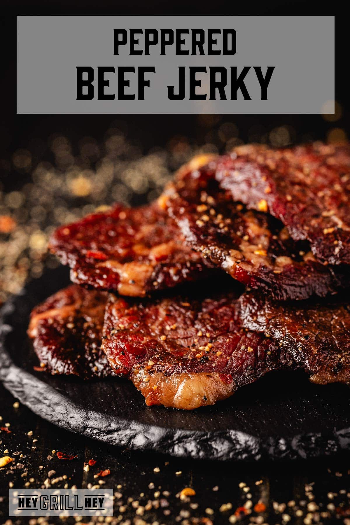 Beef jerky slices piled on a black platter. The text overlay reads "Peppered Beef Jerky" at the top, and "Hey Grill Hey" at the bottom.