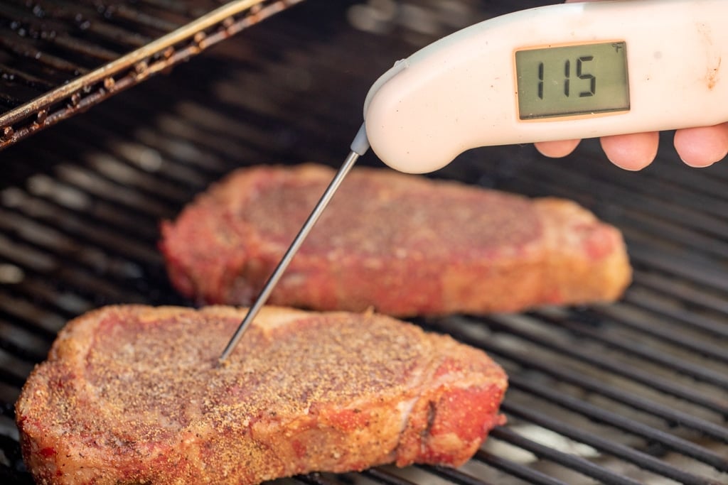 white meat thermometer taking the temperature of a seasoned steak on a smoker with the temperature reading 115 degrees F.