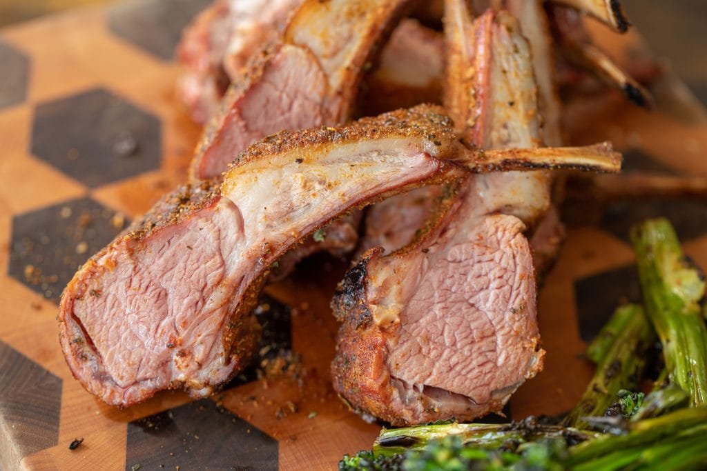 Sliced and stacked rack of lamb next to some grilled brocolli on a wood cutting board.
