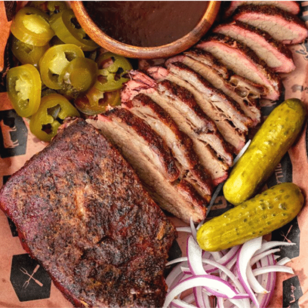 Tri tip cooked like a brisket sliced and arranged on peach butcher paper next to pickles, onions, BBQ sauce, and sliced jalapenos.