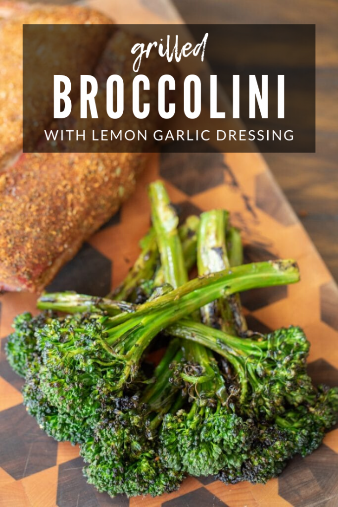 Stack of grilled broccolini and seasoned meat on a wooden cutting board with text overlay: "Grilled broccolini with lemon garlic dressing."