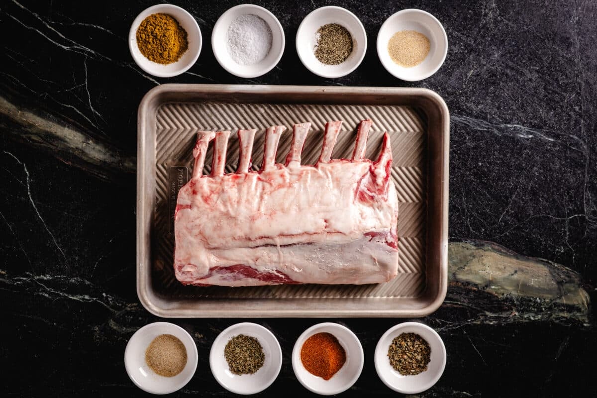 Raw meat on a baking sheet surrounded by 8 bowls with individual ingredients for a seasoning blend.