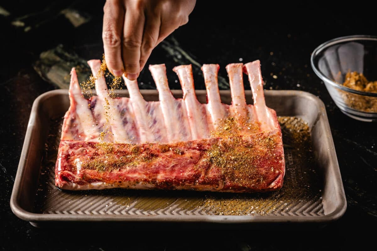 A chef's hand sprinkling a spic rub onto a rack of raw meant on a baking pan.