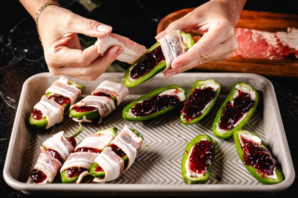 Hands wrapping a stuffed jalapeño in a slice of bacon above a baking sheet.