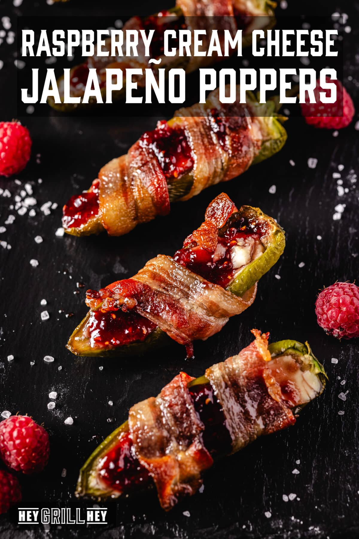 Jalapeno Poppers Three bacon wrapped jalapeños on a black platter garnished with flaky salt and fresh berries. The text overlay reads "Raspberry Cream Cheese Jalapeño Poppers" at the top, and "Hey Grill Hey" at the bottom.
