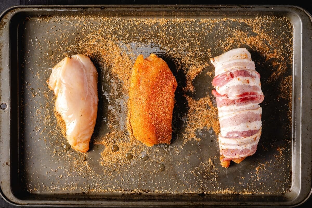Three cuts of chicken on a baking sheet. One raw, one seasoned, and one with a bacon weave.