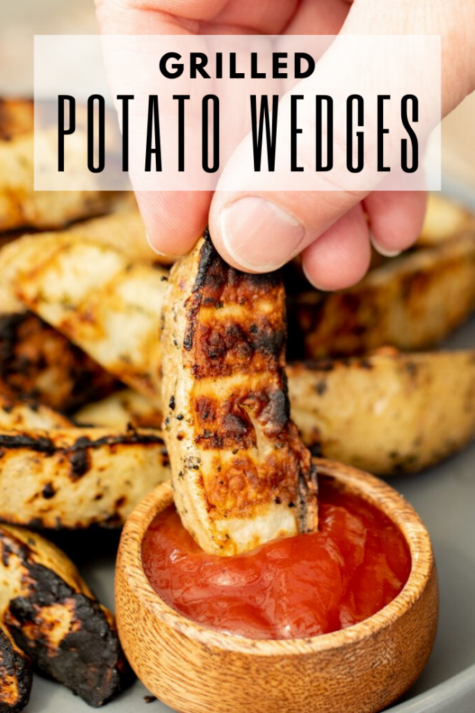 Hand dipping a grilled potato wedge into a small bowl of ketchup with a pile of grilled potato wedges in the background. Text overlay: "Grilled Potato Wedges."
