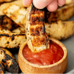 Single grilled potato wedge being dipped into ketchup with a pile of grilled potato wedges in the background.