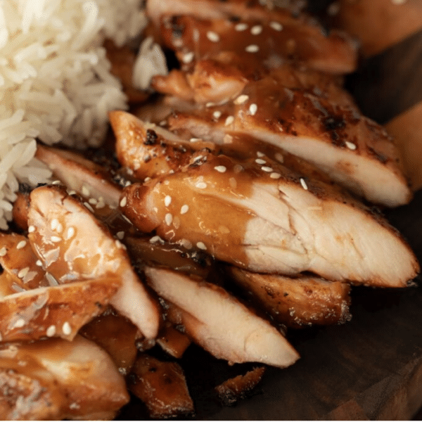 Sliced teriyaki chicken arranged on a wood cutting board and topped with teriyaki sauce.