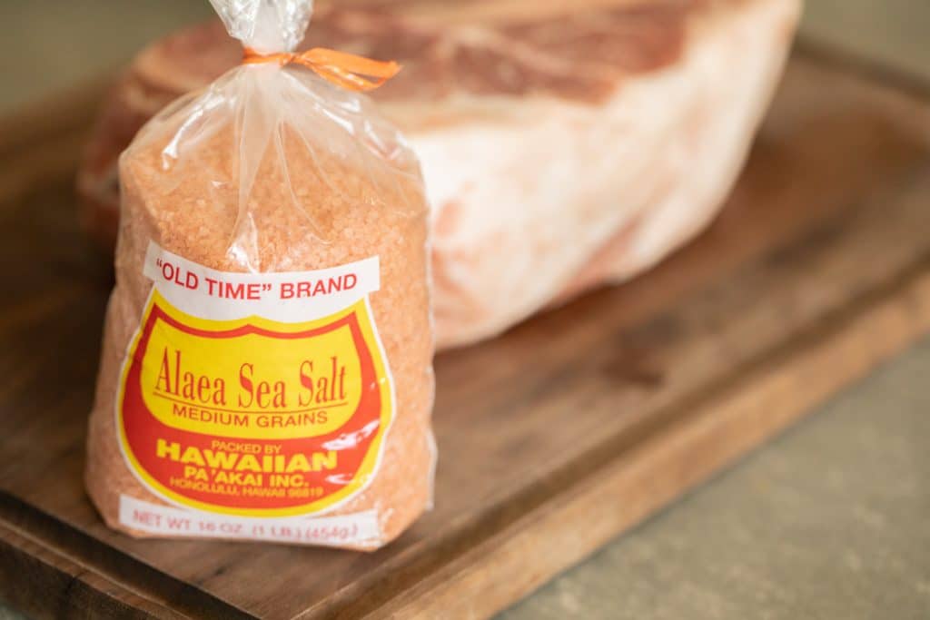 A bag of pink Hawaiian salt on a wood cutting board, with a raw pork shoulder in the background