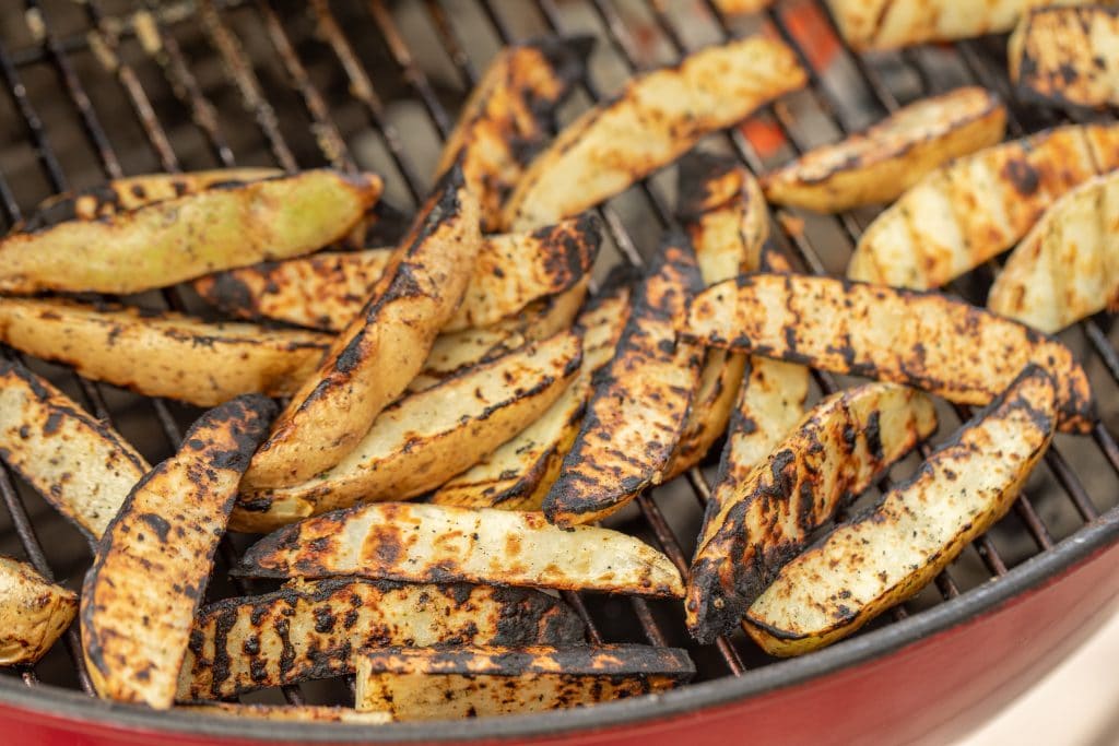 Grilled potato wedges on the grill grates of a charcoal grill.