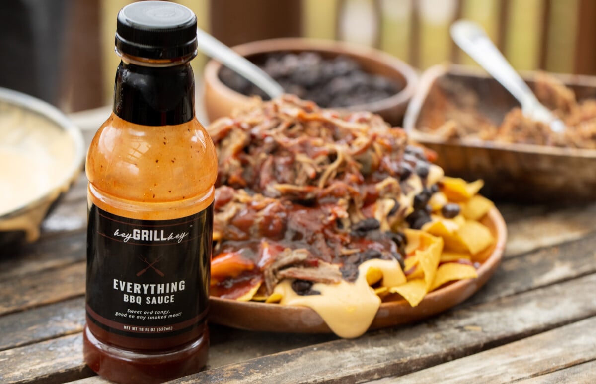 Bottle of Hey Grill Hey Everything Sauce in the foreground with a plate of pulled pork nachos in the background.