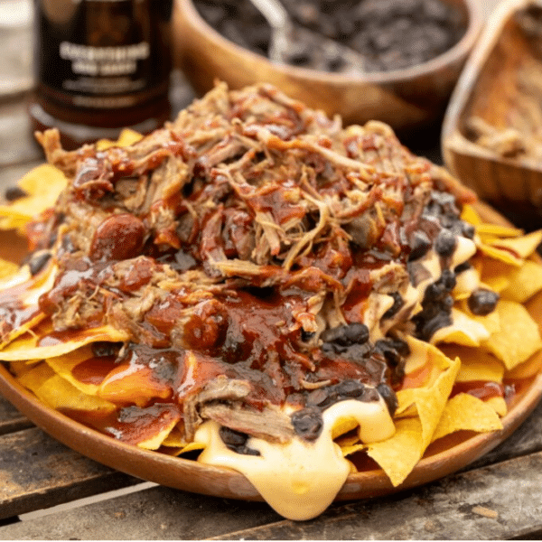 Pulled pork nachos on a wooden plate with a bottle of Everything Sauce and bowl of black beans in the background.