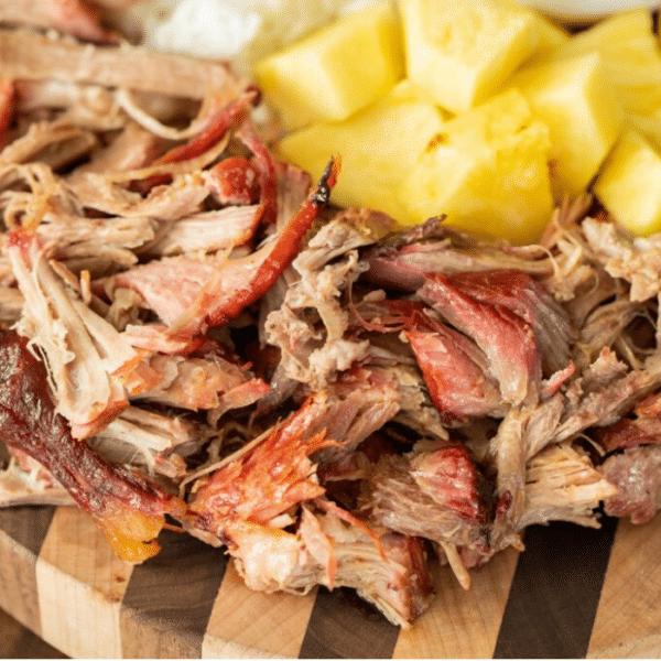 Shredded kalua pork on a wood cutting board with pineapple chunks in the background.