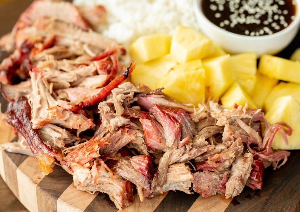 Shredded kalua pork on a wood cutting board in the foreground with pineapple chunks, rice, and a small bowl of teriyaki sauce in the background