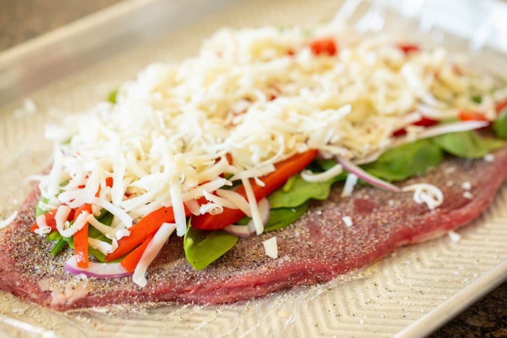 Flattened flank steak on a piece of plastic wrap sitting on a metal baking sheet. Flank steak is seasoned and topped with uncooked spinach, roasted bell pepper strips, red onions, and a pile of shredded mozzarella cheese.