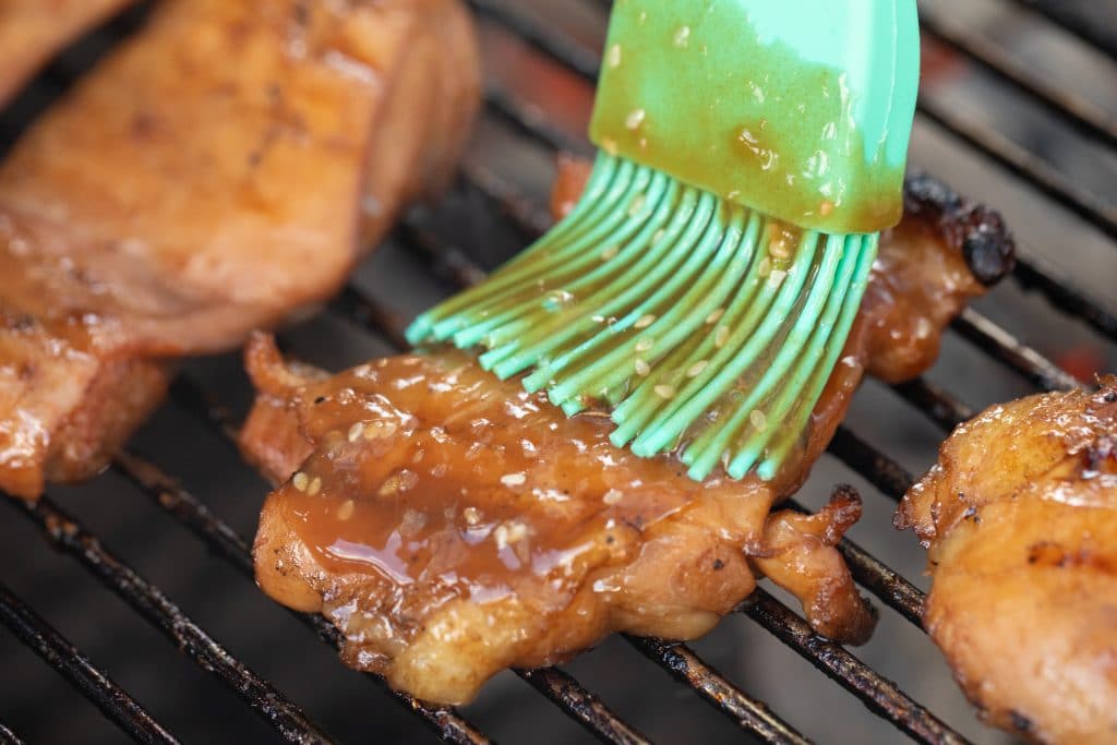 Blue colored basting brush, basting teriyaki sauce onto a chicken thigh on a grill