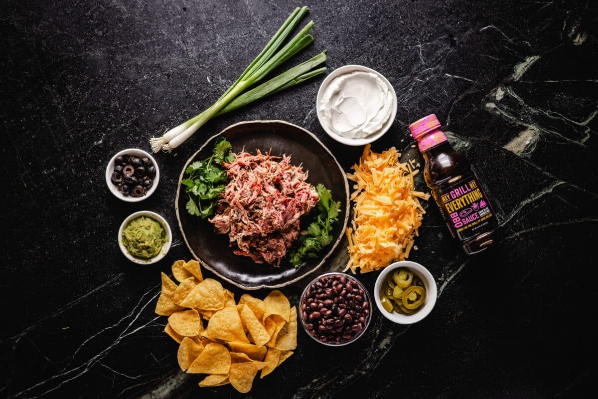 An overhead shot of chips, smoked meat, and other toppings for nachos.