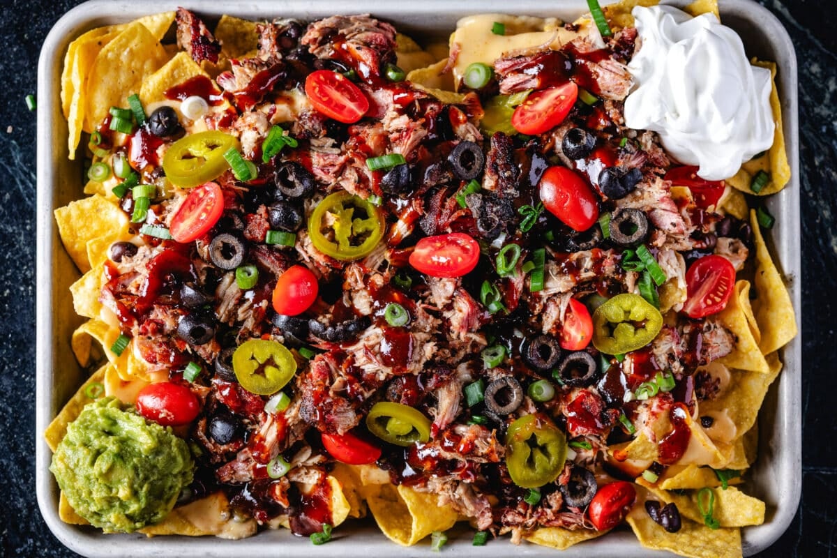 A baking sheet of nachos topped with pulled pork and tomatoes, and garnished with dollops of guacamole and sour cream.