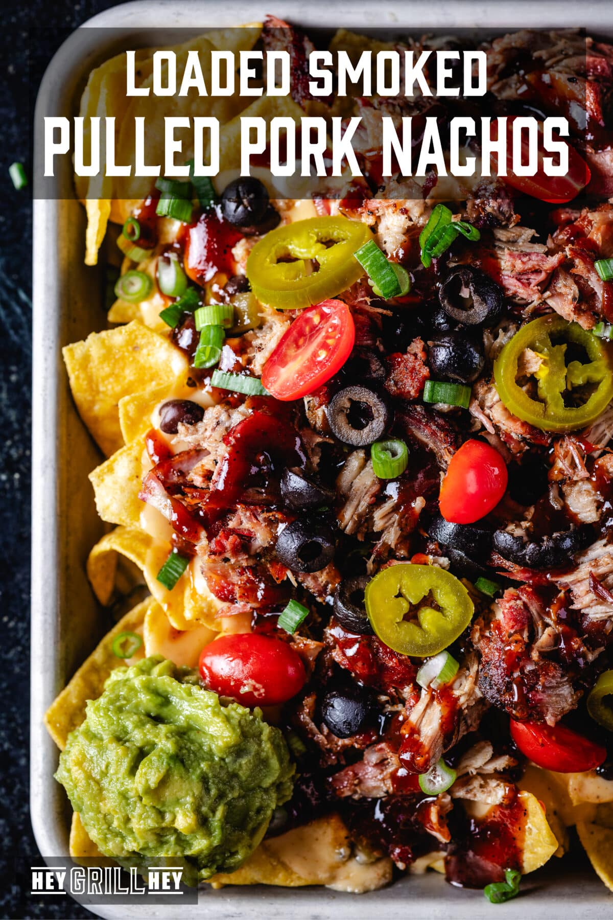 An overhead shot of a tray of nachos topped with tomatoes, jalapeños, and pork. The text overlay reads "Loaded Smoked Pulled Pork Nachos" at the top, and "Hey Grill Hey" at the bottom.