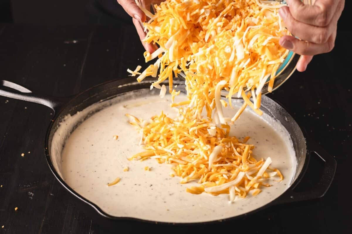 Shredded cheese being added to a cheese sauce in a cast-iron skillet.