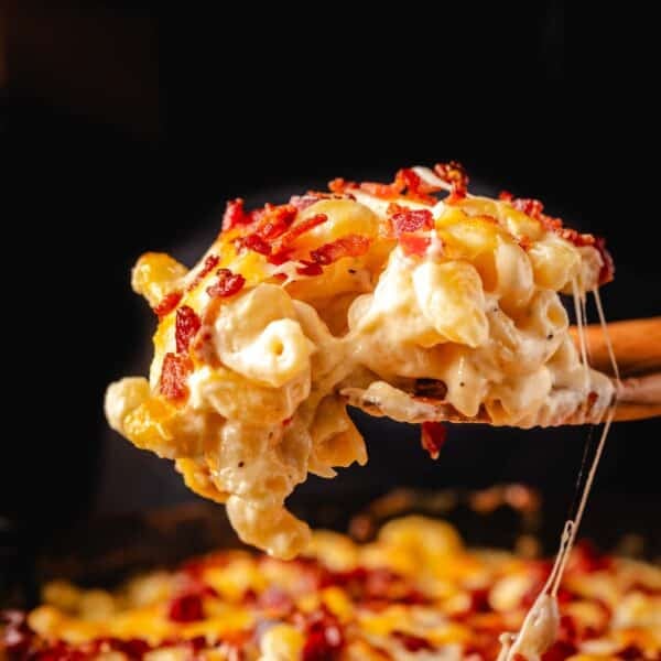 A wooden spatula holding a scoop of bacon mac and cheese over a skillet.