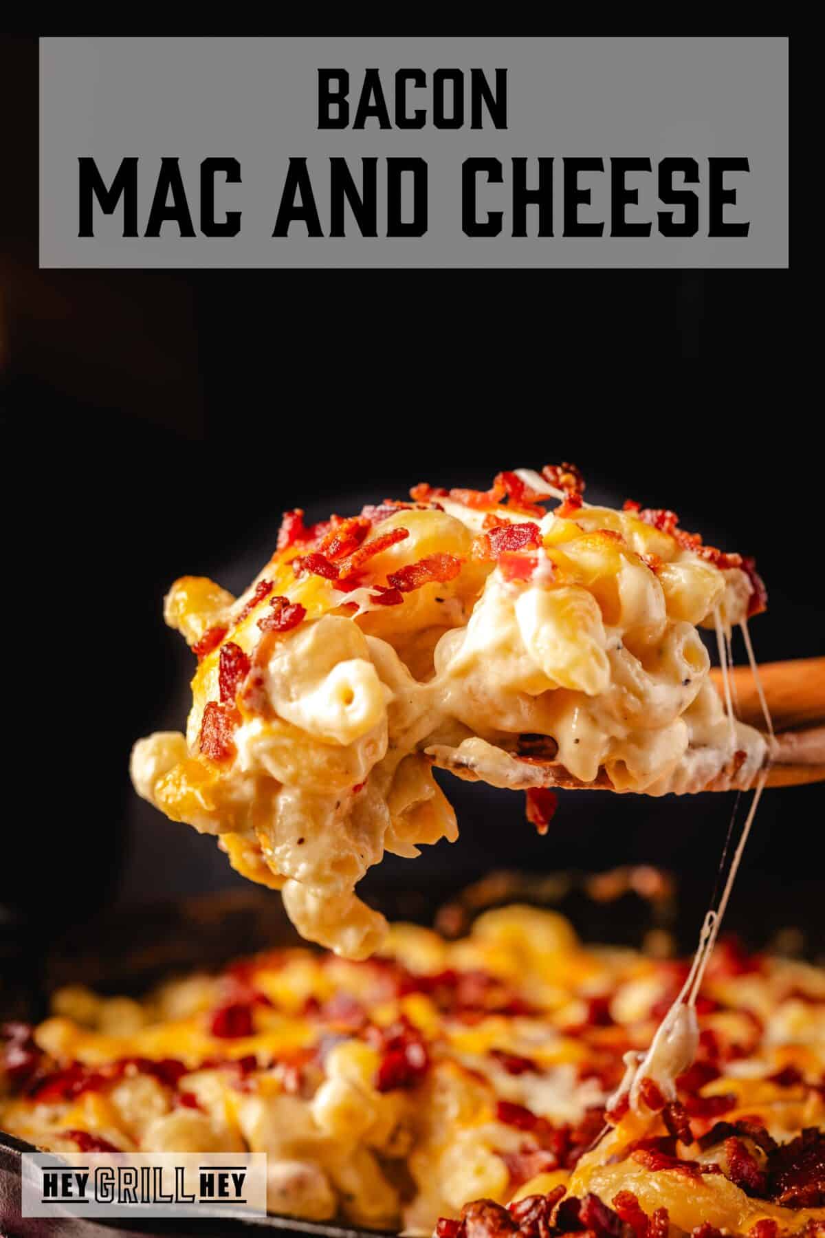 A wooden spatula holding a scoop of macaroni over a skillet. The text overlay reads "Bacon Mac and Cheese" at the top, and "Hey Grill Hey" at the bottom.