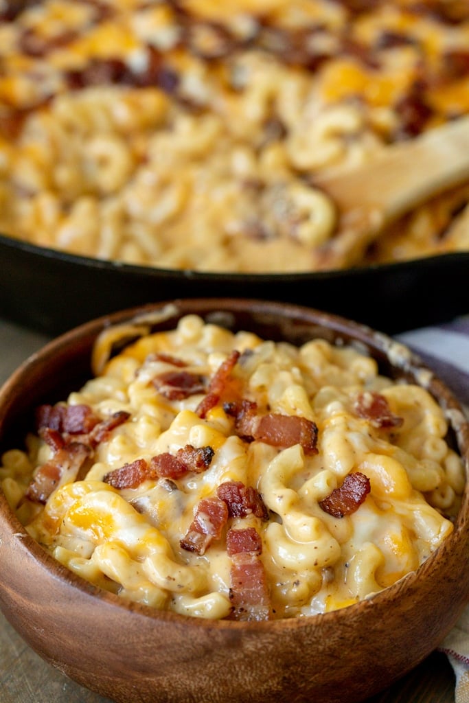 Bacon mac and cheese in a wooden bowl with a skillet of bacon mac and cheese in the background.