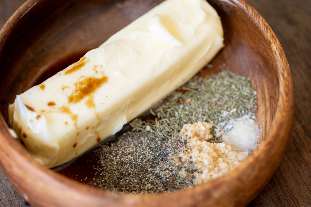 A stick of butter in a wooden bowl with brown liquid sauce and various herbs and seasonings.