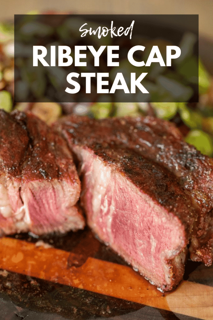 Sliced Ribeye Cap Steak with a portion turned and showing edge to edge medium rare. Grilled brussels sprouts in the background, all on a wood cutting board. Text Overlay: Smoked Ribeye Cap Steak.