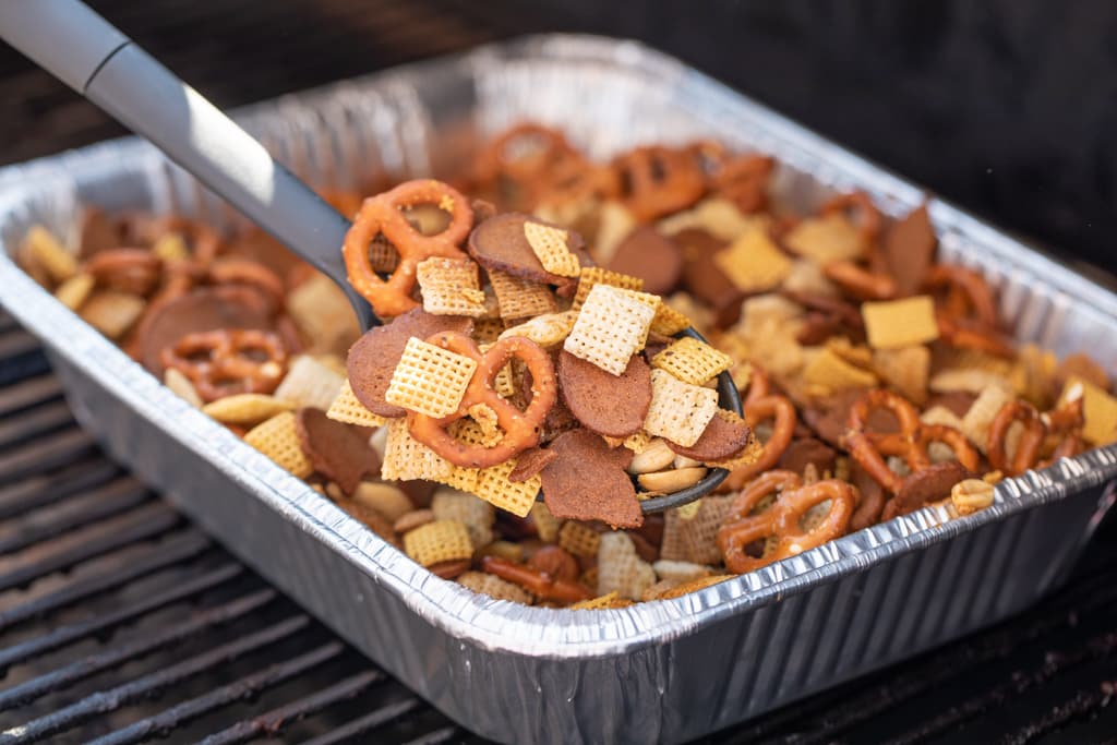 Spoonful of smoked chex mix over an aluminum pan of smoked chex mix on the grill grates of a smoker.