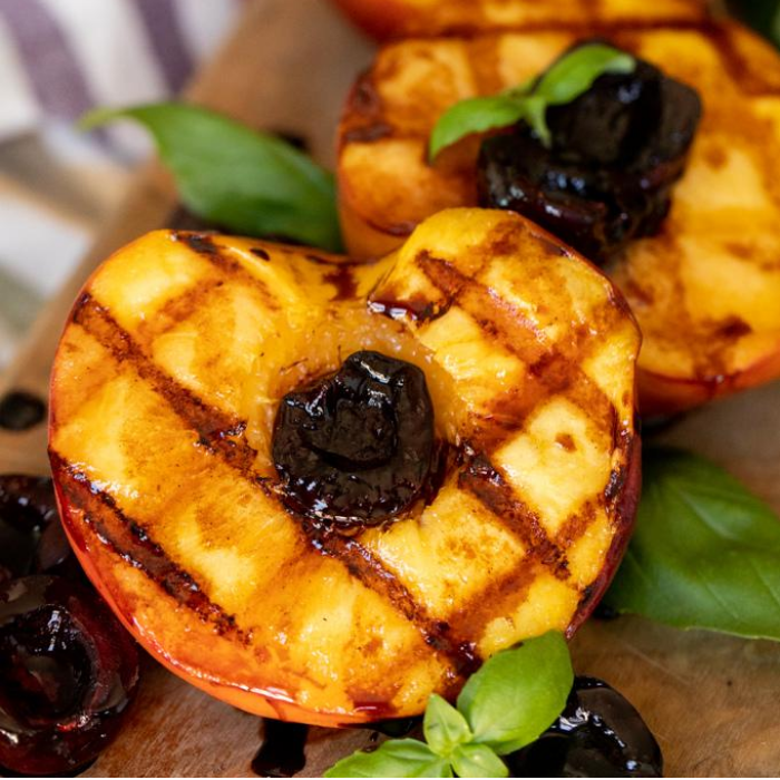 Two grilled peaches on a wooden cutting board garnished with fresh basil and dark cherries.