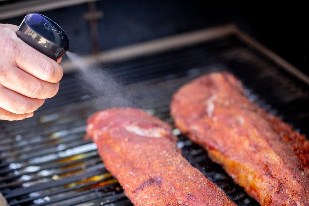 Spray bottle being squeezed and liquid misting on to the smoking ribs below.