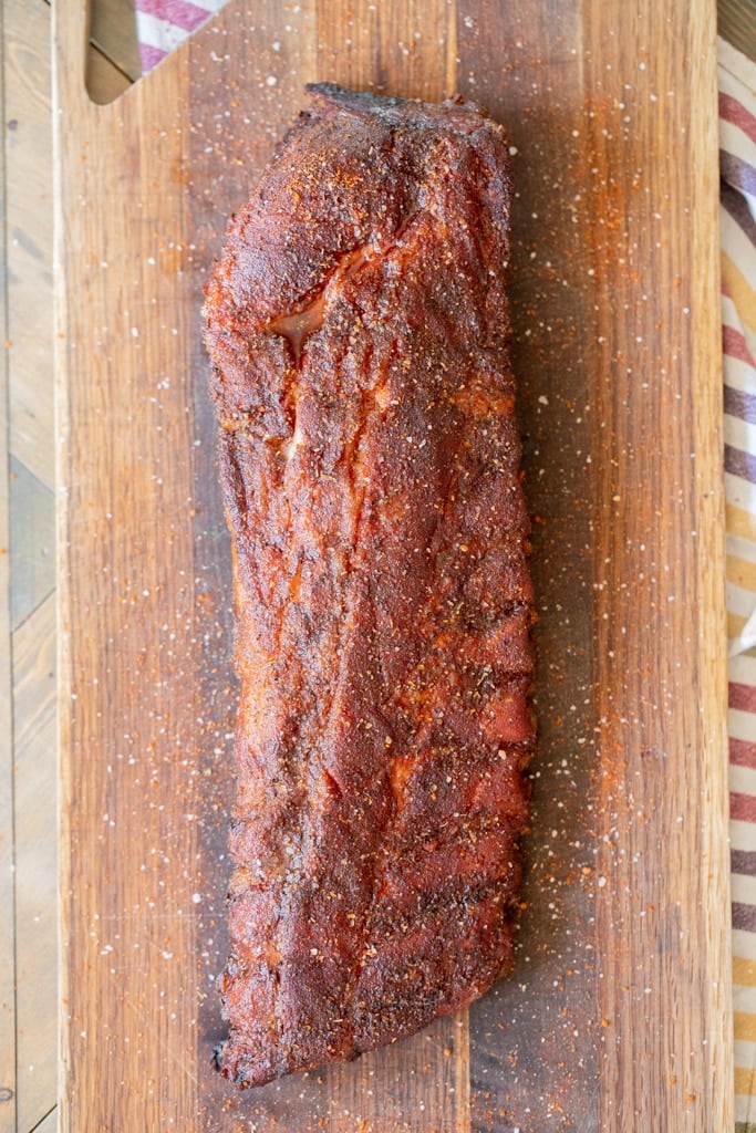Overhead view of a rack of smoked baby back ribs on a wood cutting board. Seasoning has been sprinkled on top of the ribs.