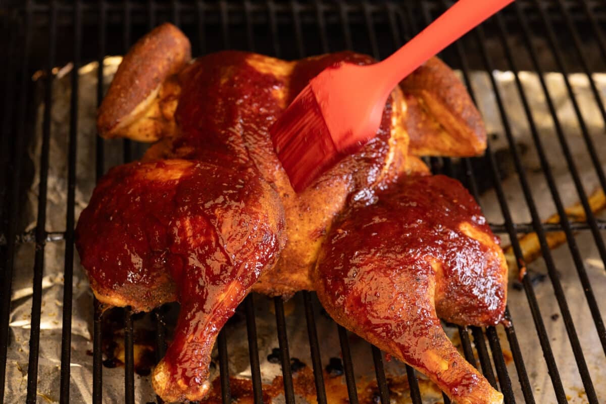 A brush basting a spatchcock chicken sitting on the grates of a smoker.