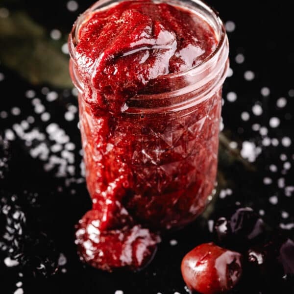 A glass jar overflowing with red sauce onto a salted counter topped with a cherry.