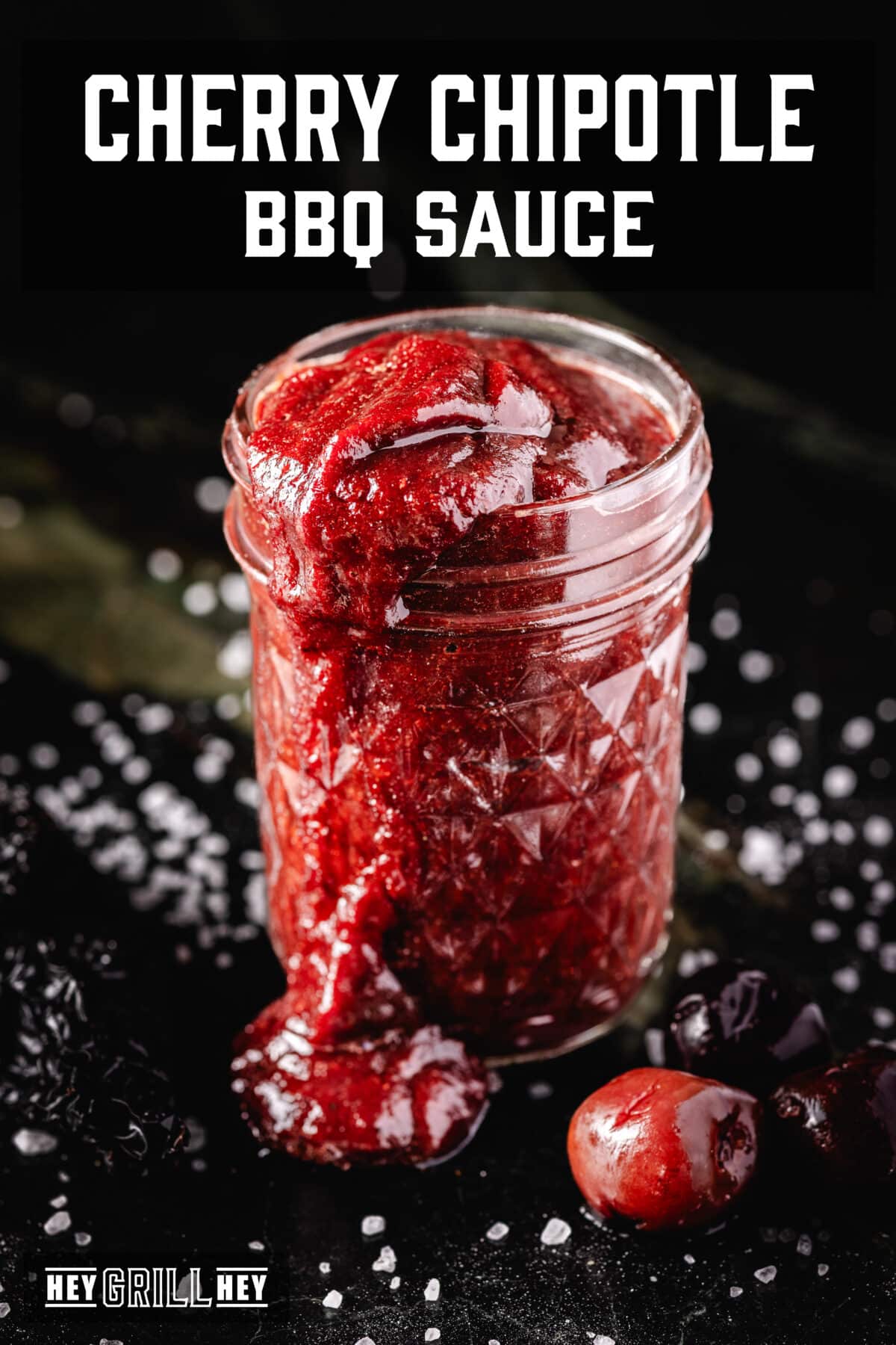 A glass jar overflowing with red sauce onto a salted counter topped with a cherry. The text overlay reads "Cherry Chipotle BBQ Sauce" at the top, and "Hey Grill Hey" at the bottom.