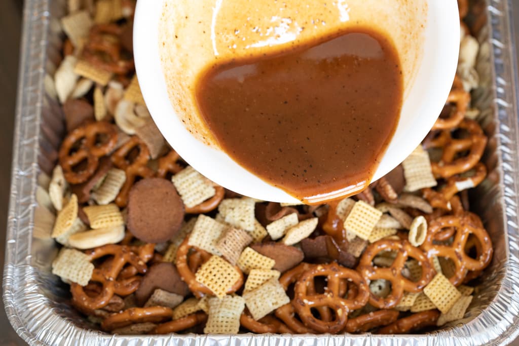 White bowl of melted butter, Worcestershire sauce, and Sweet Rub being poured over Chex snack mix.