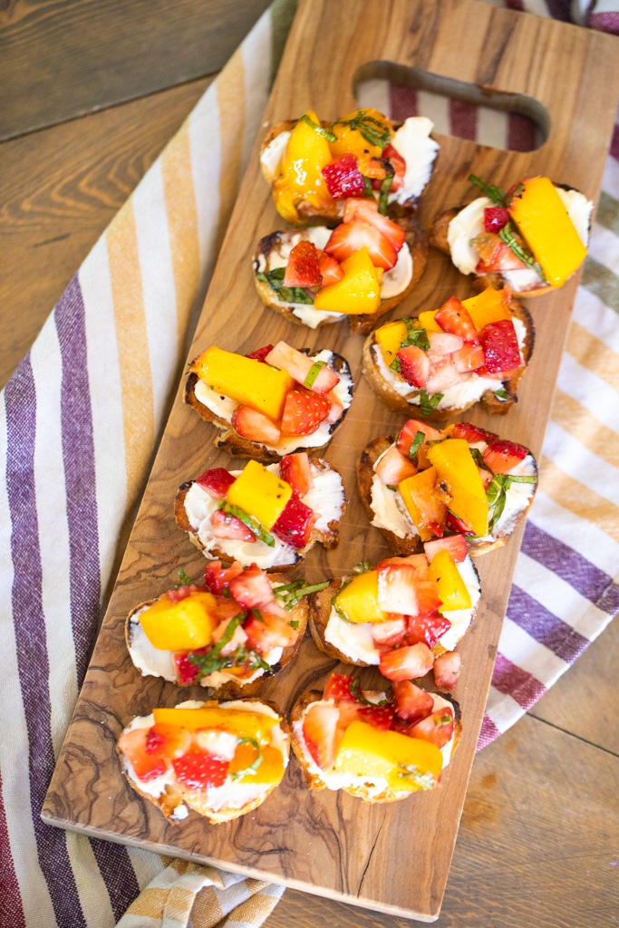 Toasted small bread rounds, topped with sweetened mascarpone and diced strawberries and peaches on a wooden serving board set on a striped dish towel.