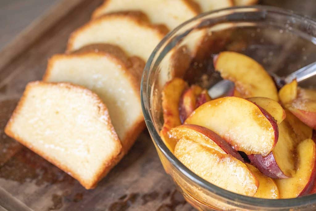 Glass bowl of sliced peaches next to a line of sliced pound cake on a wooden cutting board.